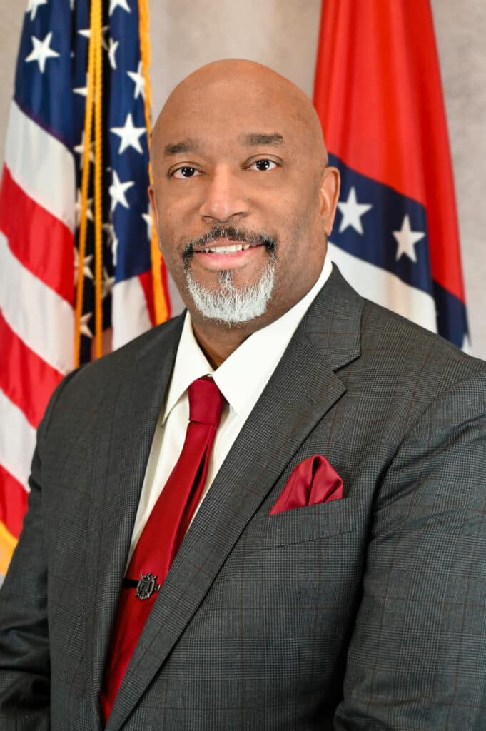 Arkansas Department of Corrections Secretary Dexter Payne, wearing a grey suit and smiling for his portrait in front of the United States and Arkansas flags