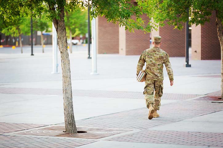 A male in a military uniform walking towards a school building carrying text books, past green trees.