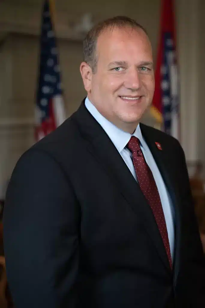 Energy & Environment Secretary Shane Khoury, smiling for his portrait in a dark suit, standing in front of the US flag