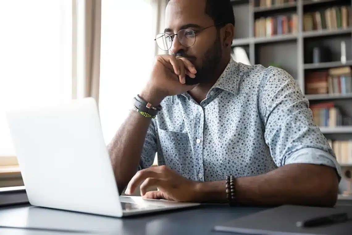 Thoughtful millennial biracial man in eyeglasses stack with hard task, looking at laptop screen. Puzzled young african ethnic businessman thinking of problem solution, making difficult decision.