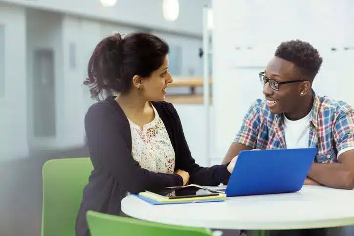 A female teacher and male student sitting at a round table with a blue laptop sitting in front of them discussing how to fund his education going forward.