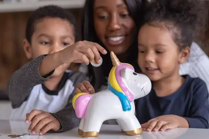 A beautiful mother of African American descent is teaching her two children of elementary school age the importance of saving money for a brighter future. The adorable children are adding all of their pocket money to the unicorn shape money box.