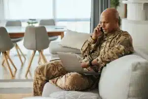 An African American Military solider in uniform sitting on his couch talking on the phone while looking at a laptop.
