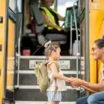 Mother with her daughter who is wearing a green backpack at a school bus stop, about to jump on the yellow school bus to go to school.
