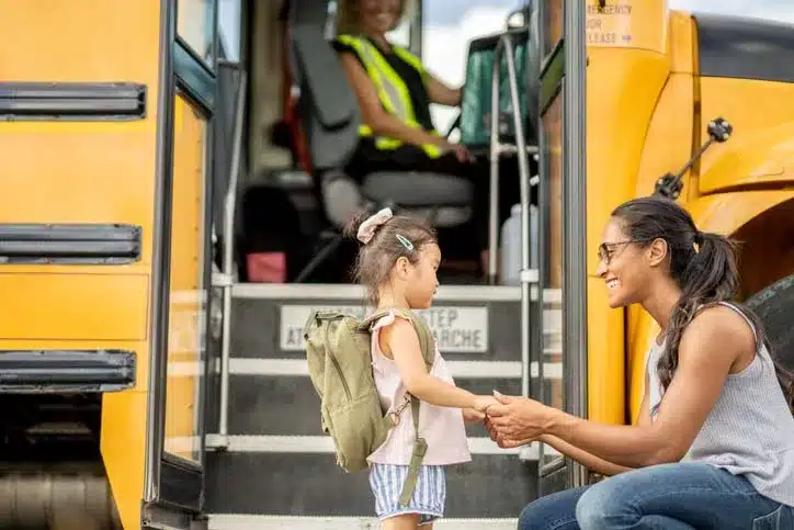 Mother with her daughter who is wearing a green backpack at a school bus stop, about to jump on the yellow school bus to go to school.