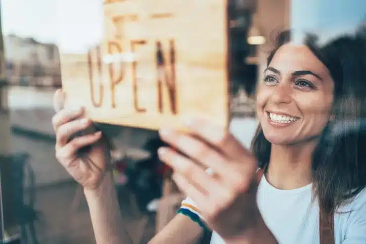 A smiling female in a short sleeve shirt working at a coffee shop flipping the wooden open sign to open.