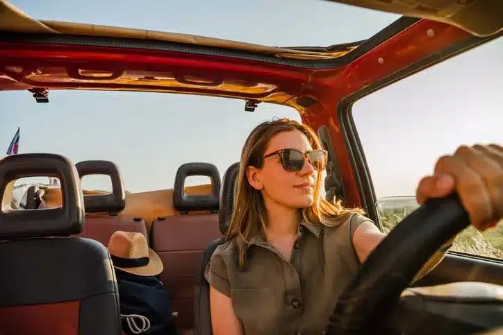 A single female wearing sunglasses driving a red vehicle with the sunroof open and the windows down looking into the sunset.