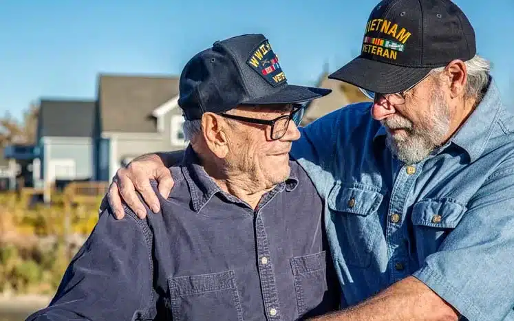 Two elderly male veterans hugging wearing a Vietnam veteran cap and the other one wearing a World War II cap.